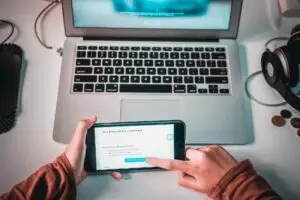 Overhead view of a laptop keyboard on a desk, with headphones and a person holding a phone showing a nomination message on screen.
