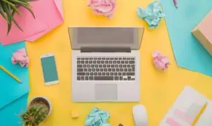 Bright workspace with a silver laptop, smartphone, notebooks, and scattered crumpled papers on a yellow surface with colorful accents.