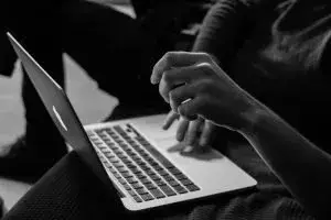 Person typing on a silver laptop resting on their lap (black-and-white photo).
