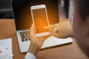 Person holding a smartphone in front of a laptop on a wooden desk with a warm orange glow on the phone screen in the foreground