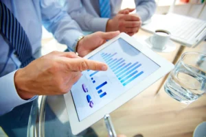 Businessperson points to a tablet displaying blue bar and line charts during a meeting.