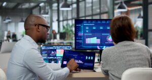 Two colleagues analyze data dashboards on multiple screens in a bright open-plan office.