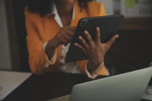 Person in an orange blazer uses a tablet at a desk with a laptop nearby, focused on the screen.