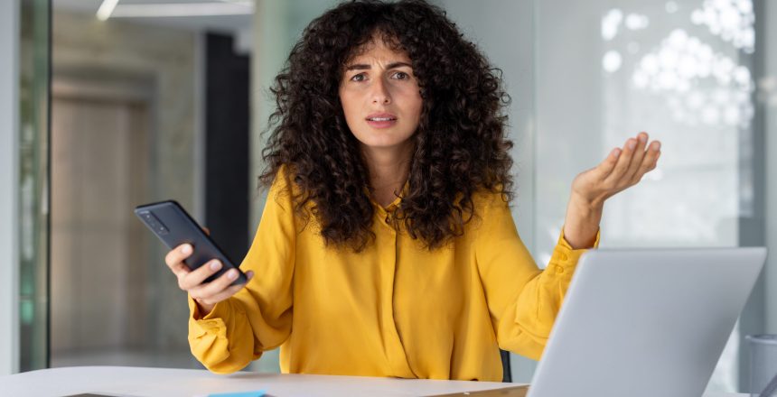 Frustrated curly haired businesswoman in yellow blouse sits at a desk holding smartphone gesturing confusedly while using laptop in modern office