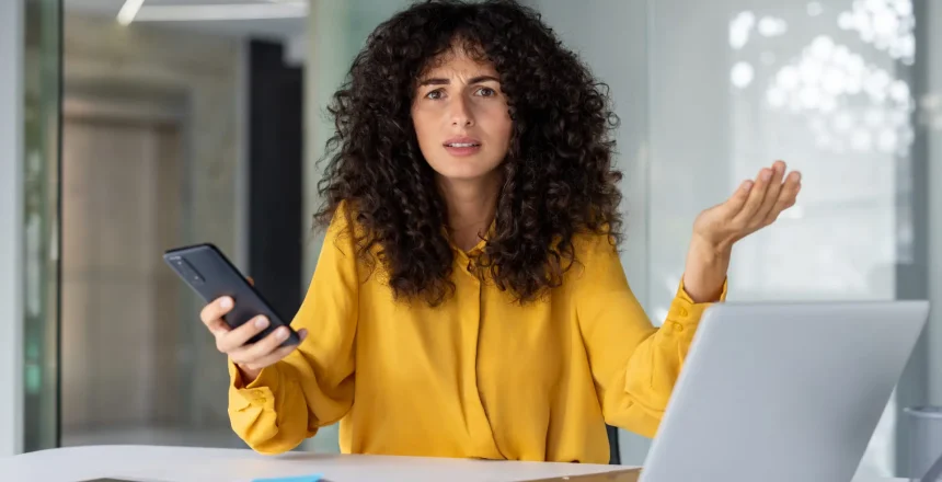 Frustrated curly haired businesswoman in yellow blouse sits at a desk holding smartphone gesturing confusedly while using laptop in modern office