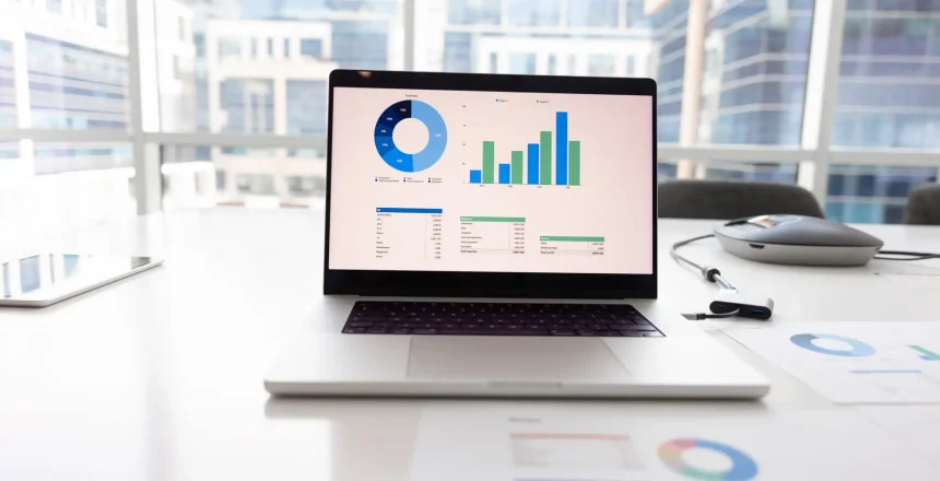 Open laptop on workplace table with analytic diagrams, financial data, statistic charts, economical infographics on screen. Marketing analyst workplace with computer and paper reports, no people