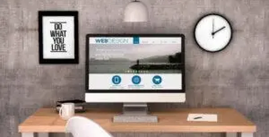 Home office desk with monitor showing a website, framed quote, round wall clock, and white chair.