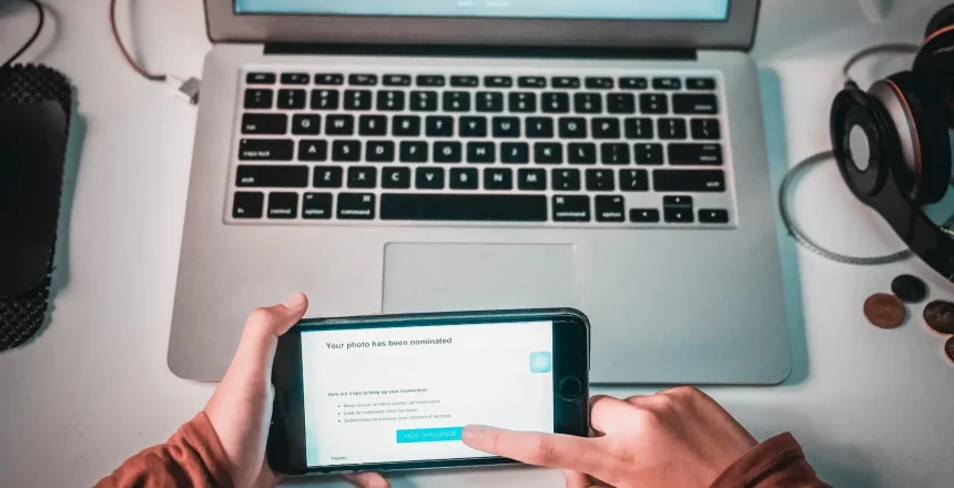 Overhead view of a laptop keyboard on a desk, with headphones and a person holding a phone showing a nomination message on screen.