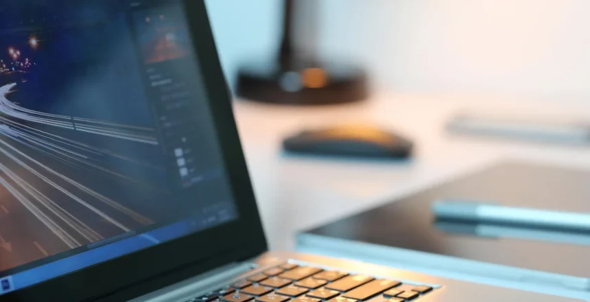 Close-up of a laptop keyboard with a blurred monitor showing code or data on a desk with a lamp in the background.