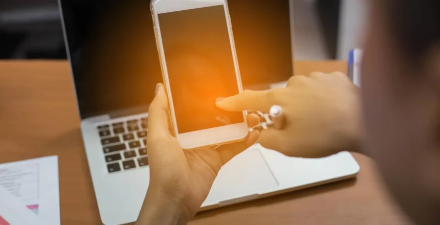 Person holding a smartphone in front of a laptop on a wooden desk with a warm orange glow on the phone screen in the foreground