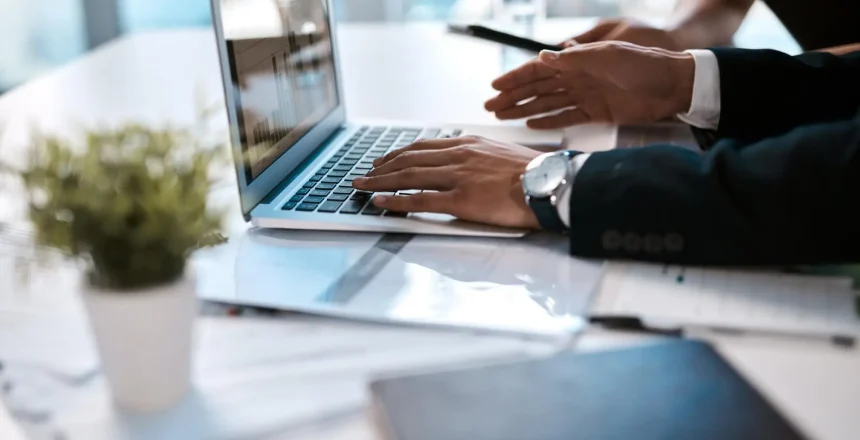 Person in a suit types on a laptop at a bright desk, with a small plant in the foreground.