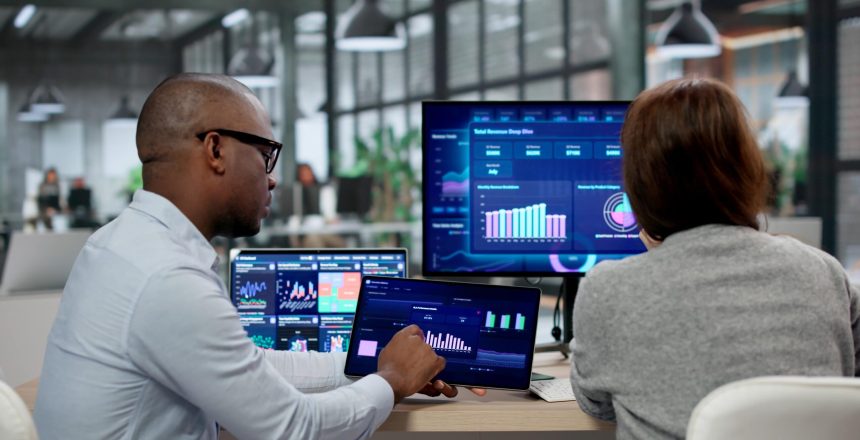 Two colleagues analyze data dashboards on multiple screens in a bright open-plan office.