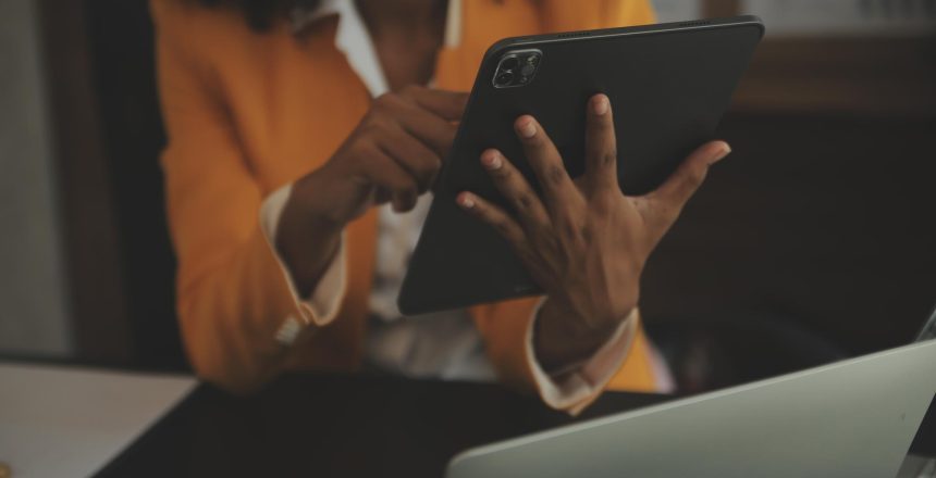 Person in an orange blazer uses a tablet at a desk with a laptop nearby, focused on the screen.